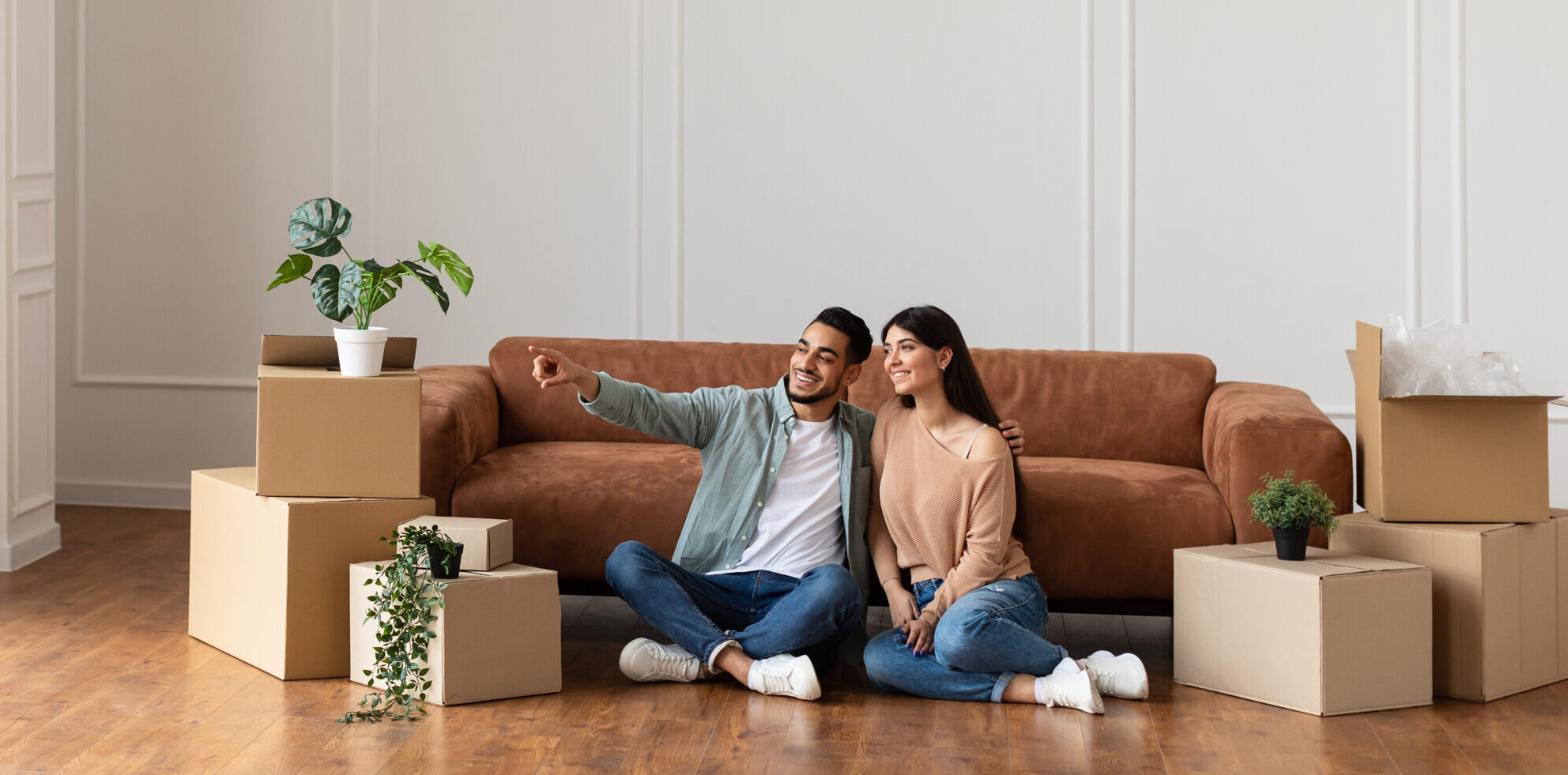 Happy man and sitting on floor choosing interior design Portrait of smiling family sitting on the floor leaning on couch in new empty apartment. Guy pointing finger aside at free space embracing wife, choosing wall color and interior design repair, banner